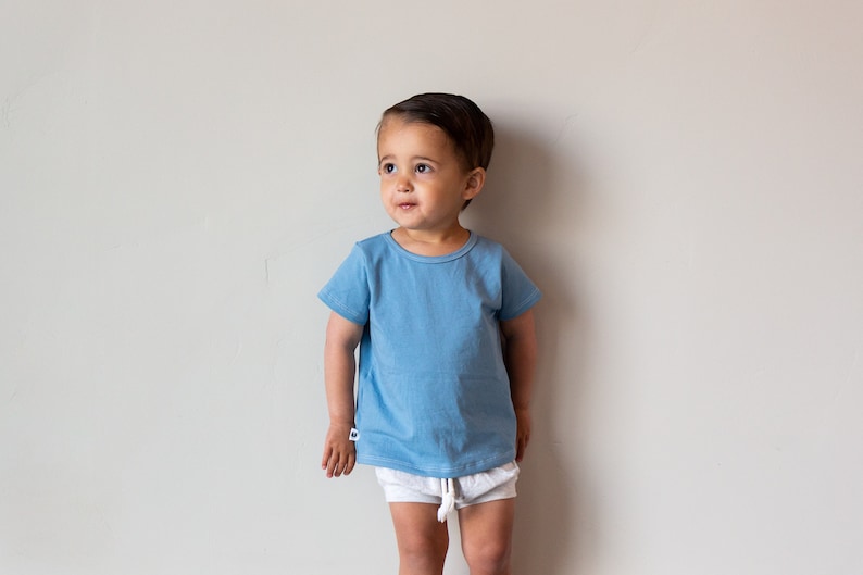 May include: A young child wearing a light blue short-sleeved t-shirt and white shorts stands in front of a white wall.