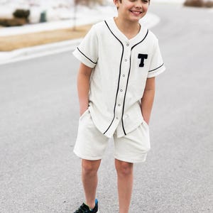 May include: A young person wearing a white baseball jersey with black trim and a black "T" on the chest. They are also wearing white shorts and black sneakers.