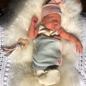May include: A newborn baby wearing a light blue and pink swaddle with a heart design and a pink knitted hat. The baby is lying on a white fluffy surface with a wooden heart-shaped teether.