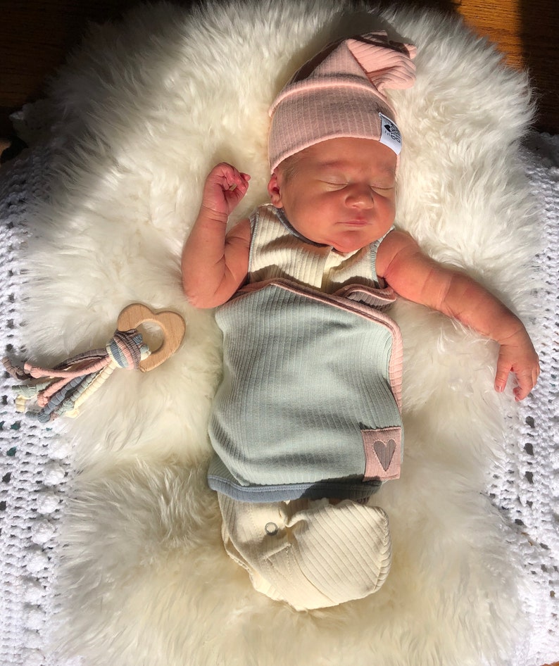 May include: A newborn baby wearing a light blue and pink swaddle with a heart design and a pink knitted hat. The baby is lying on a white fluffy surface with a wooden heart-shaped teether.