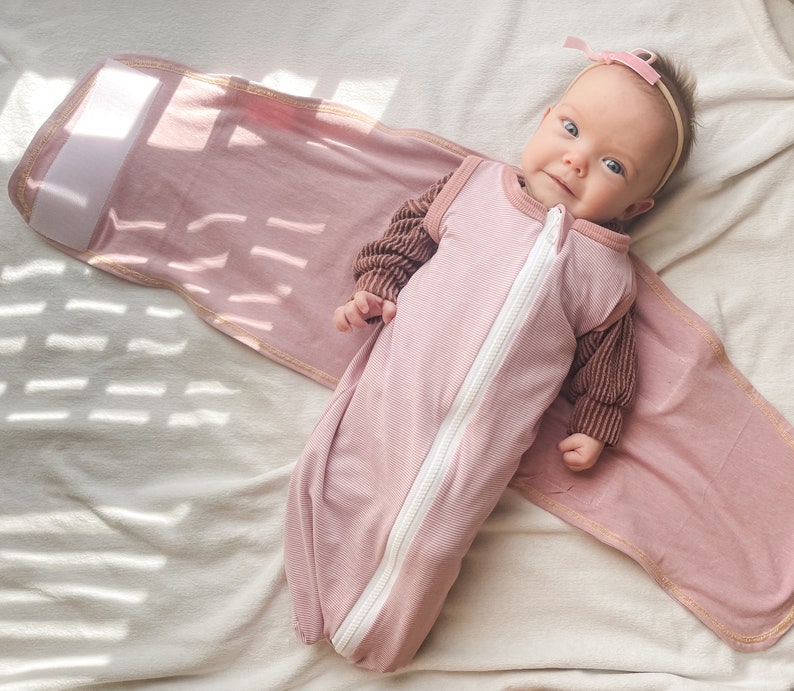 May include: A pink baby sleep sack with a zip down the front and brown sleeves. The baby is wearing a pink headband with a bow.