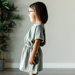 May include: A young person wearing a light green linen dress with a cinched waist and short sleeves. The dress has a relaxed fit and a simple design.