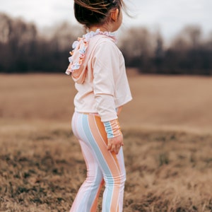Könnte beinhalten: Ein junges Mädchen trägt einen rosa Kapuzenpullover mit Rüschen und eine gestreifte Schlaghose und steht in einem Feld mit trockenem Gras.