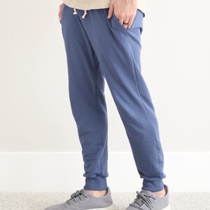 May include: A pair of blue sweatpants with a drawstring waist and cuffed ankles. The sweatpants are being worn with grey trainers.