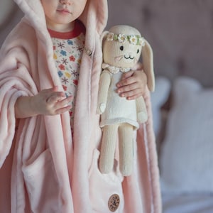 May include: A pink hooded plush robe with a pocket and a small embroidered logo. A child is wearing the robe and holding a white knitted bunny doll with a flower crown.