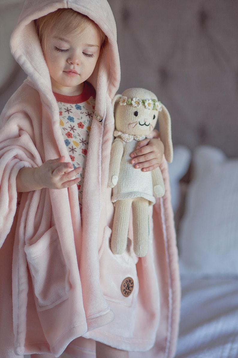 May include: A pink hooded plush robe with a pocket and a small embroidered logo. A child is wearing the robe and holding a white knitted bunny doll with a flower crown.