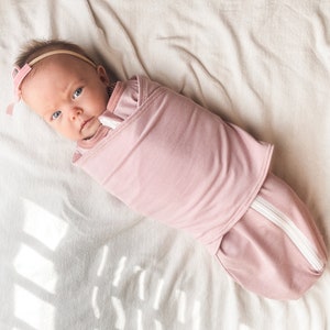 May include: A pink swaddle with a white zipper is wrapped around a baby wearing a pink headband. The baby is lying on a white blanket.