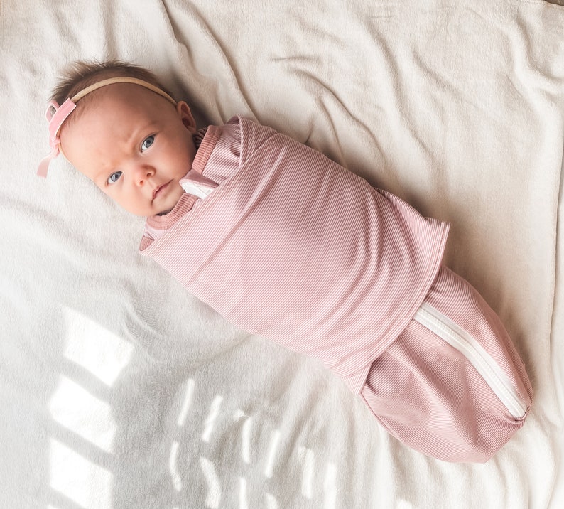 May include: A pink swaddle with a white zipper is wrapped around a baby wearing a pink headband. The baby is lying on a white blanket.