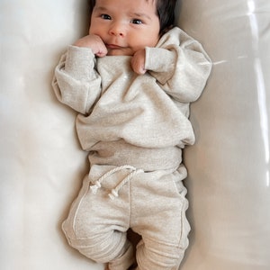 Op de afbeelding: Een baby draagt een beige sweatshirt en een bijpassende joggingbroek met een trekkoord. De baby ligt op een wit oppervlak.