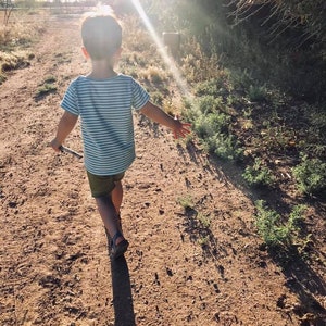 May include: A young child wearing a blue and white striped shirt and green shorts walks down a dirt path. The sun is shining brightly behind the child, creating a halo effect around their head.