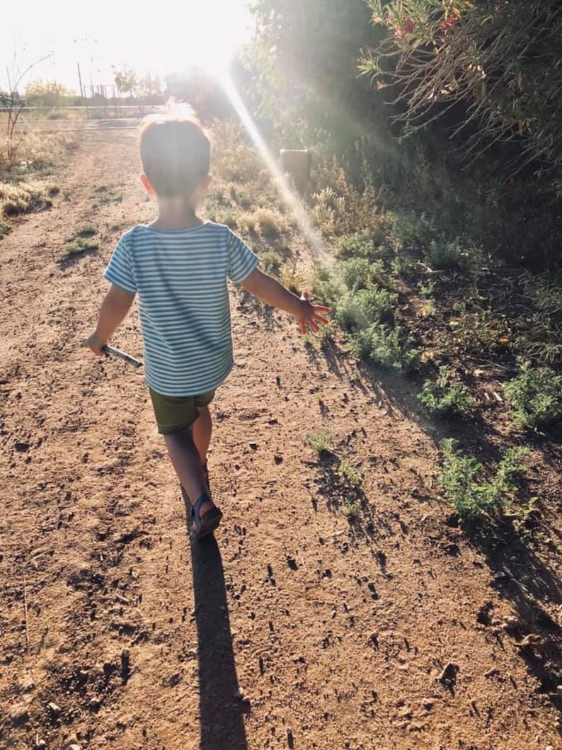 May include: A young child wearing a blue and white striped shirt and green shorts walks down a dirt path. The sun is shining brightly behind the child, creating a halo effect around their head.