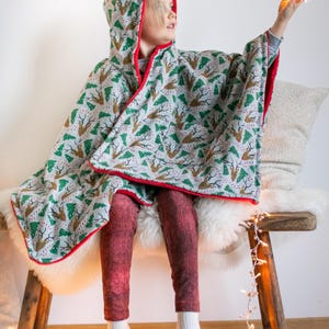May include: A child wearing a hooded, red-trimmed, green and white patterned poncho with a reindeer and Christmas tree design. The poncho is worn over red pants and white socks. The child is sitting on a wooden bench with a white furry cushion. The child is holding a white star-shaped light.