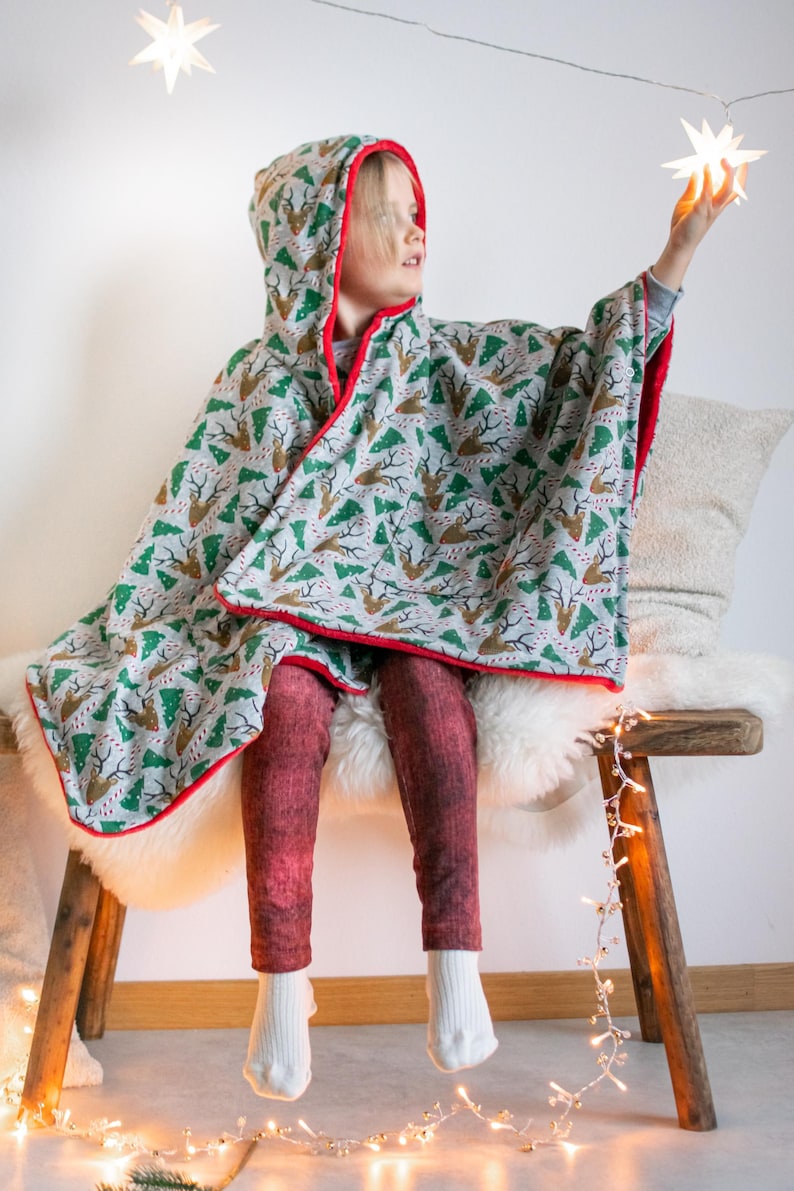 May include: A child wearing a hooded, red-trimmed, green and white patterned poncho with a reindeer and Christmas tree design. The poncho is worn over red pants and white socks. The child is sitting on a wooden bench with a white furry cushion. The child is holding a white star-shaped light.