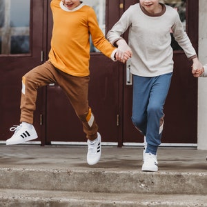 Könnte beinhalten: Zwei Kinder in langärmeligen Hemden und Hosen gehen eine Treppe hinunter. Das Kind vorne trägt ein gelbes Hemd, eine braune Hose und eine braune Baseballkappe. Das Kind hinten trägt ein beiges Hemd, eine blaue Hose und eine blaue Baseballkappe. Beide Kinder tragen weiße Turnschuhe.