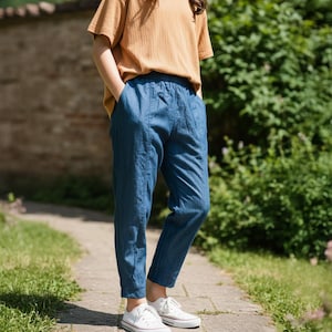 May include: A person wearing a mustard yellow short-sleeved t-shirt and blue trousers. The trousers have an elasticated waistband and are cuffed at the ankles. White trainers with red stripes complete the outfit. The person is standing on a stone path.