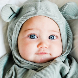 May include: A baby wearing a light green hooded onesie with bear ears. The onesie is made of a soft, ribbed fabric. The baby has blue eyes and is looking at the camera.