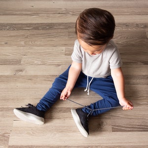 May include: A child wearing a gray t-shirt and navy blue sweatpants sits on a wooden floor, tying the laces of black and gray sneakers. The child is focused on the task.