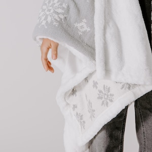 May include: A white and gray fleece blanket with a snowflake pattern. The blanket is folded and draped over a person's arm.