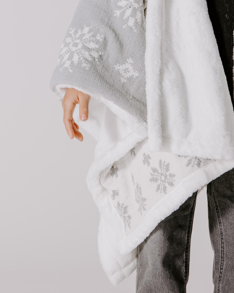 May include: A white and gray fleece blanket with a snowflake pattern. The blanket is folded and draped over a person's arm.