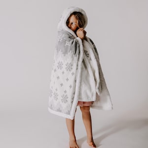 May include: A child wearing a white and gray hooded blanket with a snowflake pattern. The blanket is soft and cozy, perfect for keeping warm on a cold day.