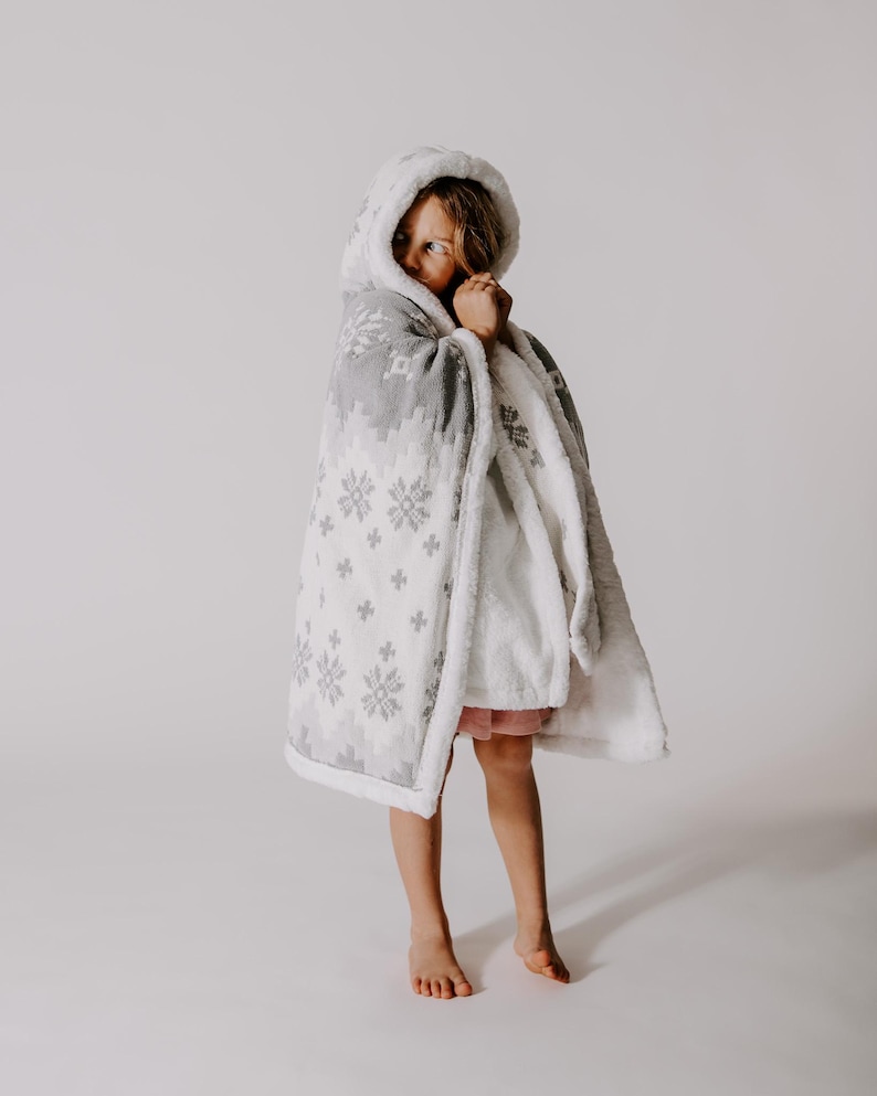 May include: A child wearing a white and gray hooded blanket with a snowflake pattern. The blanket is soft and cozy, perfect for keeping warm on a cold day.
