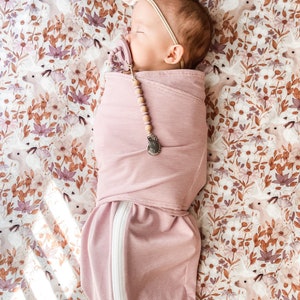 May include: A pink swaddle with white stripes and a zipper down the side. A baby is sleeping inside the swaddle. The baby is wearing a white headband and a pacifier. The swaddle is on a white and brown floral sheet.
