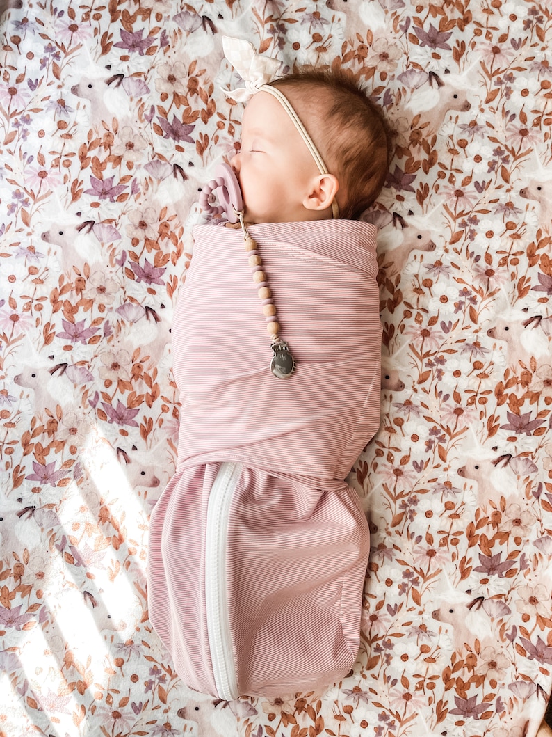 May include: A pink swaddle with white stripes and a zipper down the side. A baby is sleeping inside the swaddle. The baby is wearing a white headband and a pacifier. The swaddle is on a white and brown floral sheet.