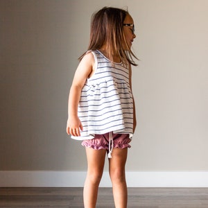 Pode incluir: Uma menina pequena vestindo um top sem mangas listrado de branco e azul com uma bainha franzada e shorts rosa franzados. Ela está em pé em frente a uma parede marrom clara.