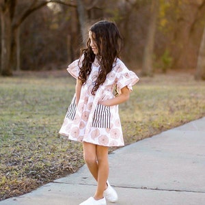 May include: A young girl wearing a white dress with a floral pattern and black and white striped pockets. She is standing on a sidewalk with green grass in the background.