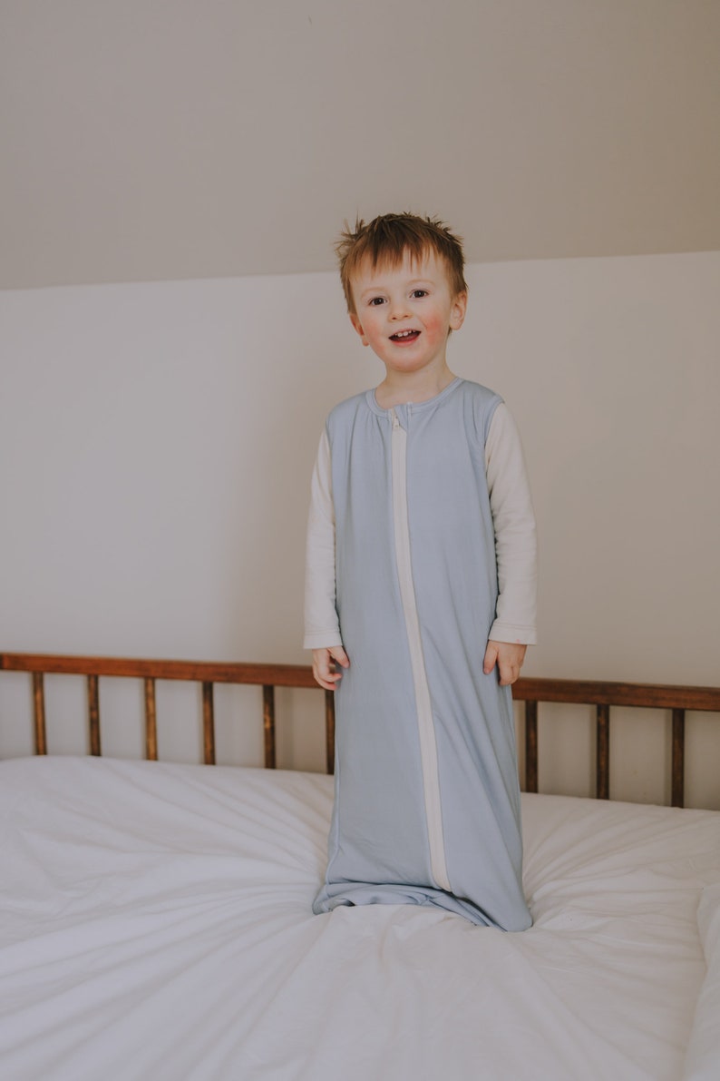 May include: A young child wearing a light blue sleep sack with a white zipper and white trim. The child is standing on a white bed with a wooden headboard.