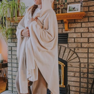 May include: A woman wearing a white hooded blanket, standing in front of a brick fireplace. She is wearing brown pants and green fuzzy slippers.