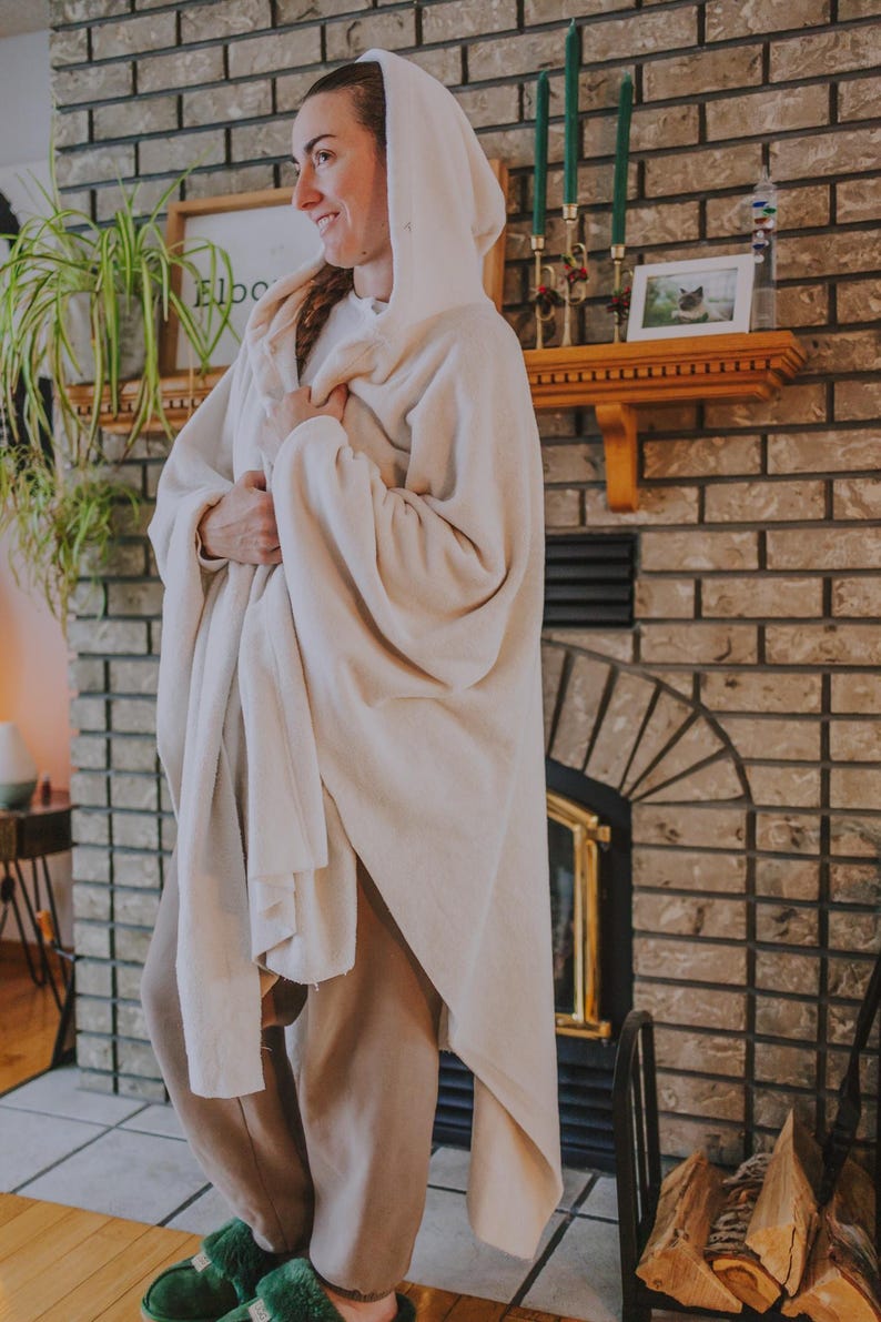 May include: A woman wearing a white hooded blanket, standing in front of a brick fireplace. She is wearing brown pants and green fuzzy slippers.