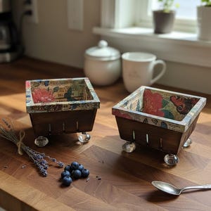May include: Two decorative wooden berry baskets with clear glass feet. The baskets have a floral and butterfly patterned interior and are displayed on a wooden surface. A bundle of lavender, blueberries, and a spoon are also present.