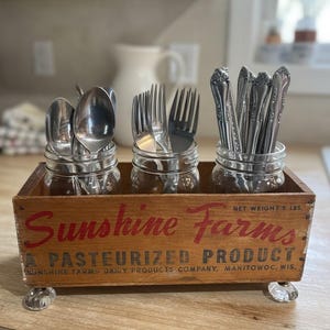 May include: A wooden utensil holder with three glass jars filled with silverware. The wooden box has the text "Sunshine Farms" in red. The box is supported by clear glass feet. The net weight is 5 lbs.