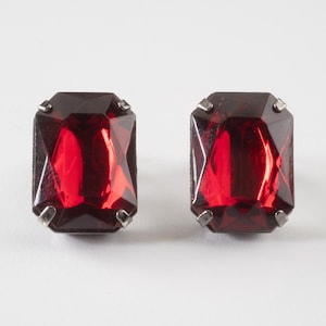 May include: A pair of rectangular stud earrings featuring deep red faceted stones. Each stone is secured by four silver-toned metal prongs at the corners, creating a classic geometric design with a polished finish and vibrant crimson color.