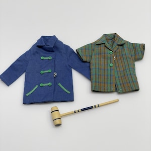 May include: A blue coat with green toggle buttons and green trim, a green and brown plaid short-sleeved shirt with green buttons, and a small wooden croquet mallet with a blue striped handle, all designed for a doll or miniature figure.