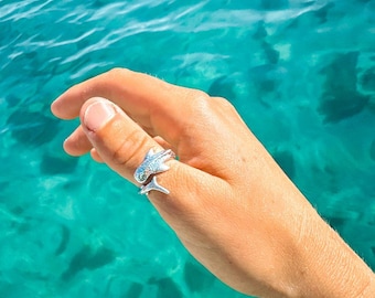 May include: A silver ring with a fish design. The ring is worn on a person's thumb. The background is a turquoise blue body of water.