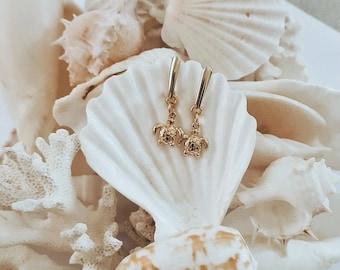 May include: Gold hoop earrings with a small turtle charm dangling from each hoop. The earrings are displayed on a white surface with seashells.