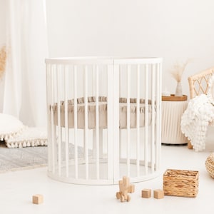 May include: A white round crib with a beige mattress and a white ruffled sheet. The crib is in a nursery with a white rug, a wicker chair, and a woven basket.