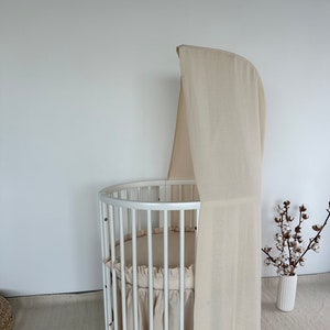 May include: White round cot with a beige canopy and a white ruffled skirt. The cot is on a beige rug and there is a vase with white cotton branches in the background.
