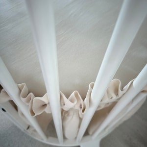 Puede incluir: Cuna de madera blanca con forro de tela blanca. La cuna tiene una forma redondeada y está hecha de listones.