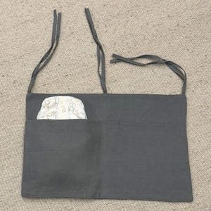 Può includere: Un organizer per pannolini in lino grigio con una tasca contenente un pannolino. L'organizer ha lunghi lacci per appenderlo. Il pannolino ha uno sfondo bianco con un motivo di animali dei cartoni animati.