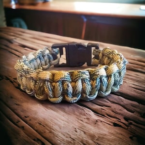 May include: A camouflage paracord bracelet with a black plastic buckle. The bracelet is made of woven paracord and has a green, brown, and yellow color scheme.