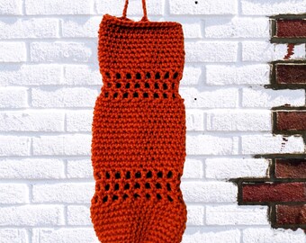 May include: A crocheted orange reusable grocery bag with a woven design. The bag has a drawstring closure and is hanging from a white brick wall.