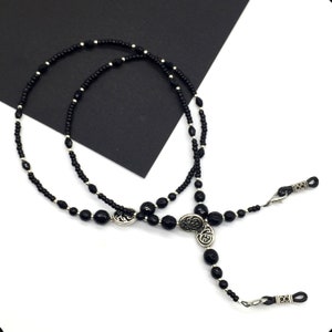 May include: Black beaded eyeglass chain with silver accents. The chain features two oval-shaped charms with a floral design.