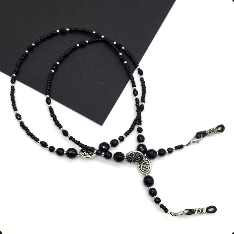 May include: Black beaded eyeglass chain with silver accents. The chain features two oval-shaped charms with a floral design.