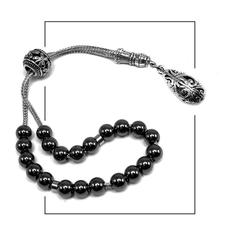 Puede incluir: Una pulsera de cuentas de oraci&oacute;n plateada con cuentas negras. Las cuentas est&aacute;n ensartadas con una cadena plateada y presentan detalles plateados decorativos, incluido un colgante en forma de l&aacute;grima. La pulsera se muestra sobre un fondo blanco.