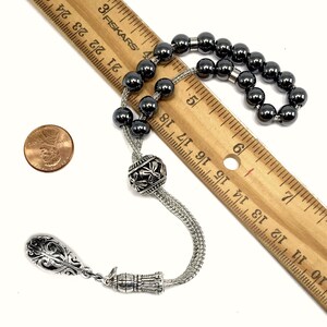 Puede incluir: Un collar de cuentas de oraci&oacute;n de color gris oscuro con un colgante decorativo plateado y una cuenta plateada con un dise&ntilde;o floral. Las cuentas tienen aproximadamente 0,8 cm de di&aacute;metro. Una moneda de un centavo y una regla est&aacute;n al lado del collar.