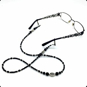 May include: Black beaded eyeglass chain with silver accents. The chain has a decorative oval charm with a floral design. The chain is attached to the glasses with silver clips.