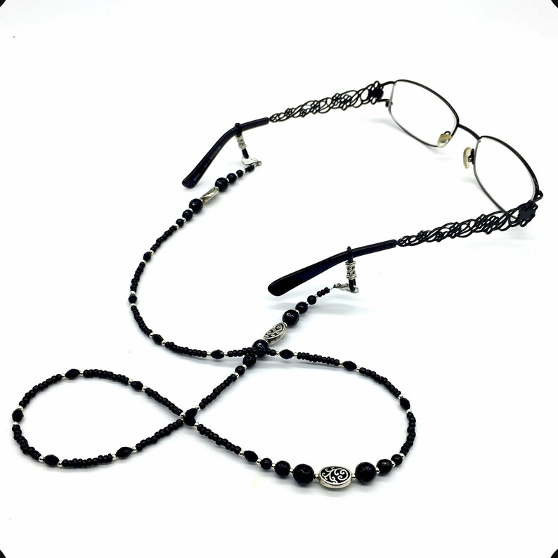 May include: Black beaded eyeglass chain with silver accents. The chain has a decorative oval charm with a floral design. The chain is attached to the glasses with silver clips.